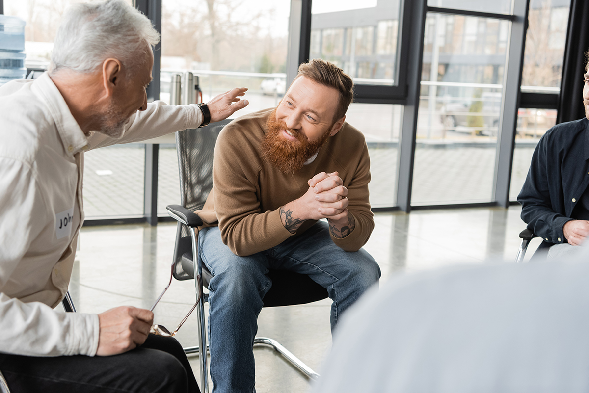 Mature man comforting smiling person with alcohol addiction in rehab center.
